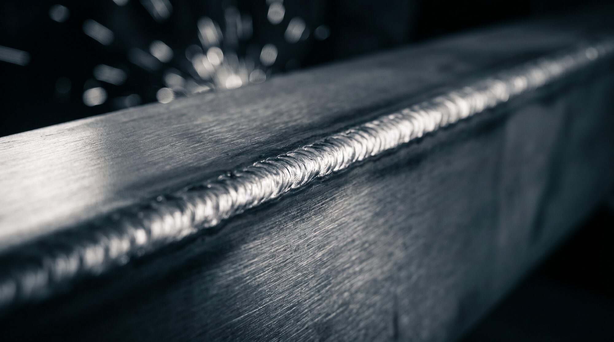 Macro close-up of a polished weld bead running along a steel trailer frame edge, sparks glowing behind